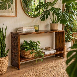 May include: A dark wood console table with a carved leaf design on the side panel. The table holds potted plants, books, and a small bowl. A round mirror hangs on the wall above the table, reflecting more plants.
