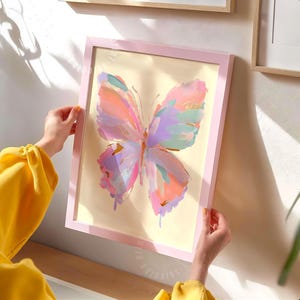 Puede incluir: Una lámina de arte enmarcada que presenta una ilustración abstracta de mariposa con pinceladas gruesas y texturizadas en tonos rosa pastel, melocotón, lavanda y verde menta sobre un fondo crema, dentro de un marco de madera rosa claro.