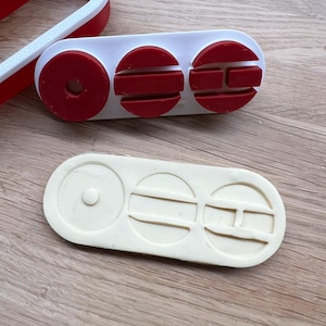 Puede incluir: Dos objetos impresos en 3D. Uno es blanco con detalles rojos, con una forma curva y una pieza rectangular con tres formas circulares y semicirculares rojas. El otro es una pieza rectangular de color beige claro con tres formas circulares y semicirculares.