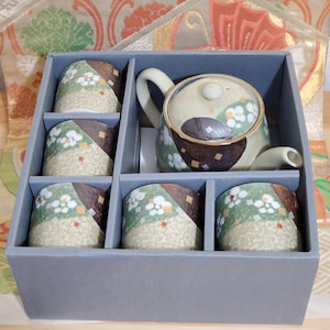 May include: A ceramic tea set with a teapot and six cups, presented in a gray box. The teapot and cups feature a cream, green, and brown floral design. The set is likely for serving tea.