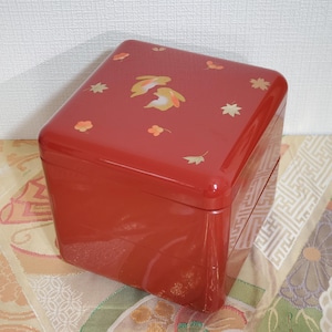 Op de afbeelding: Een rode gelakte Japanse bento-box met een vierkante vorm. Het deksel is versierd met gouden en oranje blad- en bloemontwerpen. De doos wordt waarschijnlijk gebruikt voor het vervoeren van voedsel.