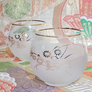 May include: Two frosted glass stemless wine glasses with gold rims. Each glass has a gold floral design. The glasses are on a patterned surface with a variety of colours.
