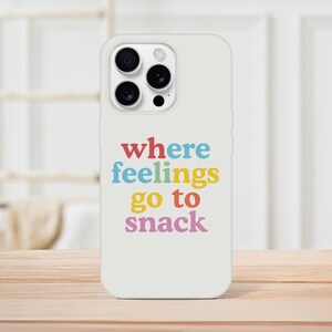 May include: A white phone case featuring the text "where feelings go to snack" in red, blue, yellow, and pink. The case is on a wooden surface, with a blurred background.