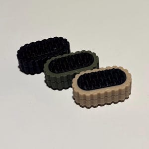 Könnte beinhalten: Drei kleine, rechteckige Objekte in Schwarz, Olivgrün und Beige. Jedes hat eine strukturierte, dunkle Mitte und einen geriffelten Rand. Die Objekte sind diagonal auf einer weißen Oberfläche angeordnet und zeigen ihre kontrastierenden Farben und Texturen.
