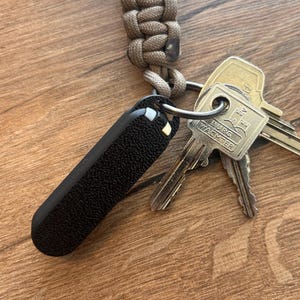 Könnte beinhalten: Ein schwarzer, strukturierter Schlüsselanhänger mit einem silbernen Ring, der Schlüssel und ein geflochtenes beiges Schlüsselband hält. Die Schlüssel enthalten einen Schlüssel mit dem Text "BURG WACHTER". Der Schlüsselanhänger liegt auf einer Holzoberfläche.