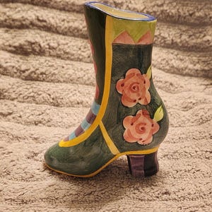 May include: A decorative ceramic boot-shaped vase. The vase is primarily dark green with colorful accents, including yellow, pink, and blue stripes. Two pink roses with green leaves are painted on the side. The heel is purple and black.