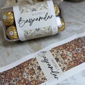 Könnte beinhalten: Eine transparente Plastikbox mit goldverpackten Schokoladen, beschriftet mit dem Text "HAYIRLI Bayramlar" in schwarzer Schrift. Das Etikett und ein Streifen darunter haben ein Muster.