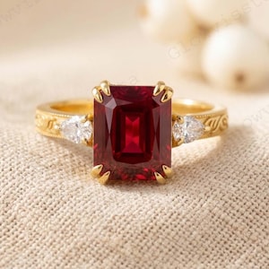 May include: A gold ring with a large, rectangular, deep red gemstone as the centerpiece. Two smaller white gemstones flank the main stone. The band has an ornate design. The ring is set against a neutral, textured background.