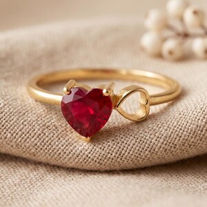 May include: A gold ring featuring a heart-shaped, deep red gemstone. The ring's band has a small heart-shaped cutout. The ring is set on a beige fabric surface, with a blurred floral element in the background.