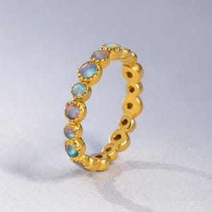 May include: A gold ring featuring a series of oval-shaped opals. The opals display a range of colors, including blue, pink, and yellow. The ring has a smooth, polished finish and is set against a light gray background.
