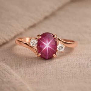 Może przedstawiać: Pierścionek z różowego złota z dużym owalnym rubinem z efektem sześcioramiennej gwiazdy. Pierścionek jest otoczony dwoma mniejszymi, okrągłymi, przezroczystymi kamieniami szlachetnymi. Pierścionek leży na tkaninie w neutralnym odcieniu.