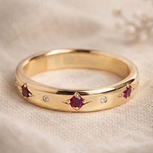 May include: A gold ring featuring red gemstones and small clear stones. The ring has a smooth, polished surface and is set with multiple red gems in a star-like setting. The ring is a piece of jewellery.