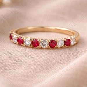 Puede incluir: Un anillo de banda de oro con piedras preciosas rojas y claras alternas. El anillo presenta pequeños rubíes de corte redondo y piedras claras, creando un diseño brillante. El anillo se muestra sobre una superficie suave de color neutro.