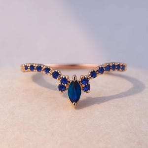 Könnte beinhalten: Ein roségoldener Ring mit geschwungenem Design, besetzt mit kleinen blauen Edelsteinen. Ein größerer, marquiseförmiger blauer Edelstein befindet sich in der Mitte. Das filigrane Design und die leuchtenden blauen Steine erzeugen eine elegante Ästhetik.