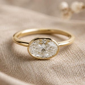 Può includere: Un anello d'oro con un grande diamante taglio ovale. Il diamante è incastonato in una montatura a castone e la fascia è in oro lucido semplice. L'anello è presentato su uno sfondo di tessuto beige.