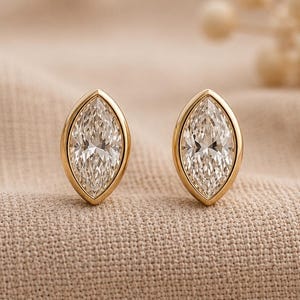 Könnte beinhalten: Ein Paar goldene Ohrstecker im Marquise-Schliff. Jeder Ohrring hat einen klaren, länglichen Diamanten, der in einer goldenen Lünette gefasst ist. Die Ohrringe werden auf einer beigefarbenen Stoffoberfläche präsentiert.
