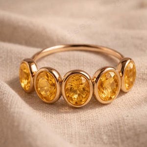 Può includere: Un anello d'oro con sei pietre preziose ovali, giallo brillante. Le pietre sono incastonate in castoni individuali lungo la fascia. L'anello è esposto su un tessuto di colore neutro.