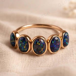 Könnte beinhalten: Ein goldener Ring mit fünf ovalen Opalen. Die Opale zeigen eine Mischung aus Blau, Grün und Rot, gefasst in einem goldenen Band. Der Ring wird auf einem neutralfarbenen Stoff präsentiert.