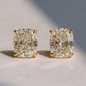 Puede incluir: Un par de pendientes de diamantes con corte cojín. Los diamantes tienen un tono champán cálido y están engastados en garras de oro. Los pendientes se muestran sobre una superficie blanca.