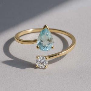 以下が含まれることがあります： ゴールドカラーのリング。 涙型のライトブルーの宝石と、丸く透明な宝石が特徴です。 リングのバンドは指に巻き付き、宝石は両端に配置されています。 リングはニュートラルな背景に配置されています。