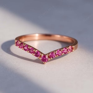May include: Rose gold V-shaped ring with a row of vibrant pink gemstones. The ring's design features a delicate curve, showcasing the sparkling gemstones. The ring is set against a soft, neutral background, highlighting its elegant simplicity.