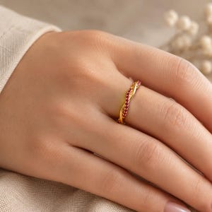 May include: A gold-colored ring with a twisted design, set with small red gemstones. The ring is on a hand with manicured nails, set against a neutral background.