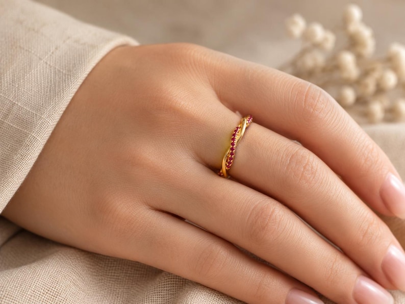 May include: A gold-colored ring with a twisted design, set with small red gemstones. The ring is on a hand with manicured nails, set against a neutral background.