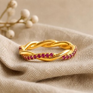 May include: A gold-colored ring with a braided design. The ring is adorned with small, vibrant pink gemstones set along the braided pattern. The ring is displayed on a neutral-toned fabric, showcasing its intricate details.