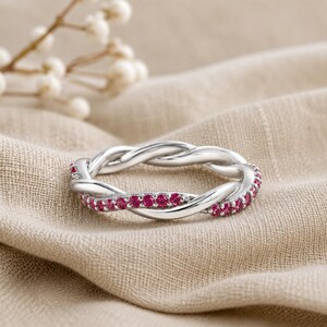 May include: A silver-colored ring with a braided design, adorned with small, vibrant red gemstones. The ring is set against a neutral-toned fabric, highlighting its intricate details and elegant style. The ring's design is a continuous loop.