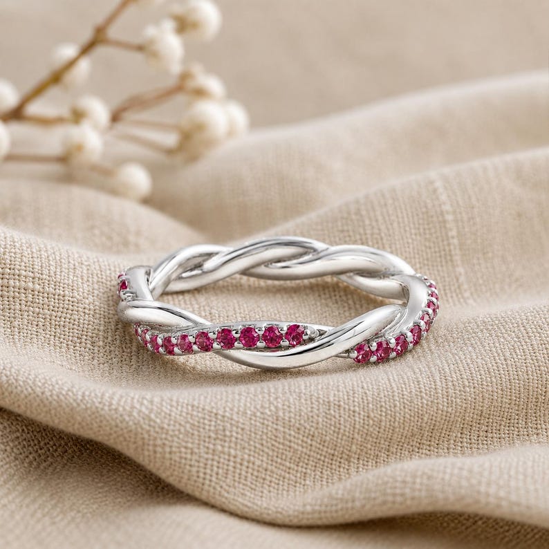 May include: A silver-colored ring with a braided design, adorned with small, vibrant red gemstones. The ring is set against a neutral-toned fabric, highlighting its intricate details and elegant style. The ring's design is a continuous loop.