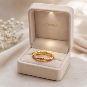 May include: A gold-colored ring with a braided design, set with small red gemstones. The ring is displayed in an open, beige velvet ring box with the text "GLOWVISSTA" embossed inside. The background is a soft, neutral fabric.