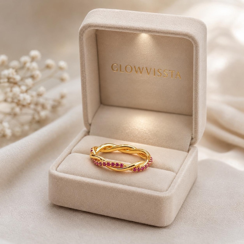 May include: A gold-colored ring with a braided design, set with small red gemstones. The ring is displayed in an open, beige velvet ring box with the text "GLOWVISSTA" embossed inside. The background is a soft, neutral fabric.