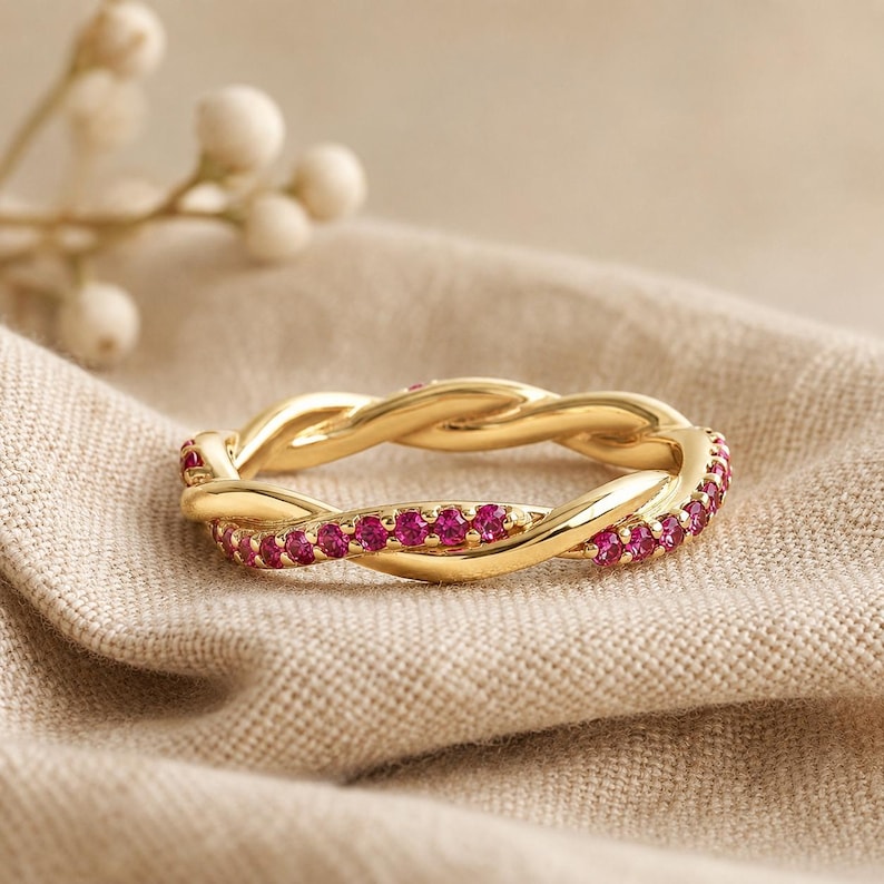 May include: A gold-colored ring with a braided design. The ring is adorned with small, vibrant pink gemstones set along the outer edges. The ring is displayed on a neutral-toned fabric.