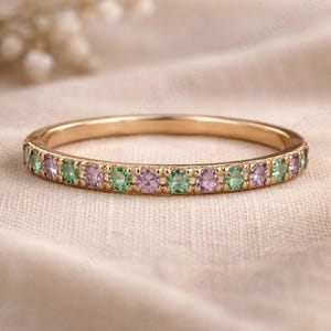 May include: A gold band ring with alternating green and purple gemstones. The ring is set with small, round gemstones that encircle the entire band. The ring is displayed on a neutral-toned surface.