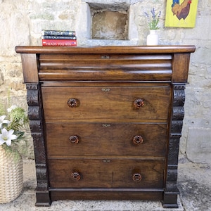 May include: A vintage wooden chest of drawers with three drawers and decorative carvings. The dresser features amber-coloured knobs and is set against a stone wall. Books are stacked on top.