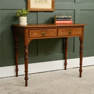 May include: A rectangular wooden writing desk featuring two drawers with brass pulls and turned legs. The desk has a warm brown wood grain finish and is topped with a small potted green plant and a stack of three books with colourful spines.