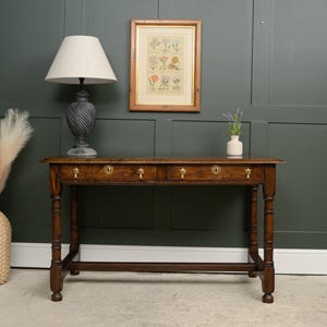 May include: An antique wooden console table with three drawers and brass handles. A framed botanical print is above the table. A gray lamp with a white shade sits on the table. A small white vase with flowers is also on the table.