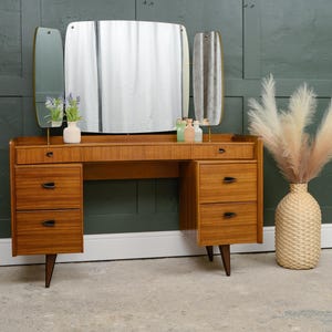 May include: A wooden dressing table with a light brown finish, featuring a central rectangular mirror flanked by two smaller adjustable side mirrors. The unit includes four drawers with black handles and a long top drawer. A woven vase with pampas grass sits nearby.