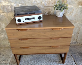 Puede incluir: Una cómoda de madera de estilo moderno de mediados de siglo con tres cajones y un tocadiscos incorporado. La cómoda tiene un tono de madera cálido con molduras marrones oscuras y patas anguladas. Una pequeña planta en maceta encima.