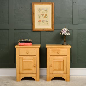 May include: Two light-toned wooden bedside cabinets, each featuring a single top drawer with a round knob and a lower cupboard door with a square recessed panel. The cabinets have a rectangular top surface and a base with arched cutouts at the bottom.