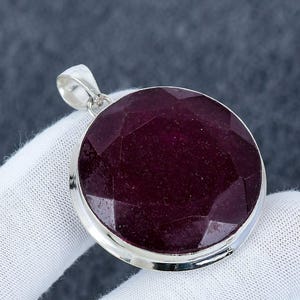 Pode incluir: Um pingente redondo de rubi vermelho escuro, facetado, emoldurado em uma moldura de prata. A pedra preciosa é o ponto focal, mostrando sua cor rica e corte geométrico. O pingente tem um design simples e elegante.