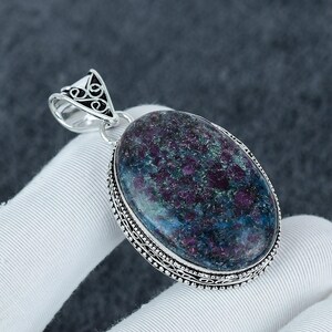 Puede incluir: Colgante ovalado con una gran piedra preciosa multicolor. La piedra presenta una mezcla de tonos morados, azules y verdes profundos. El colgante está engastado en un metal plateado con un diseño ornamentado alrededor de la piedra y una fianza decorativa.