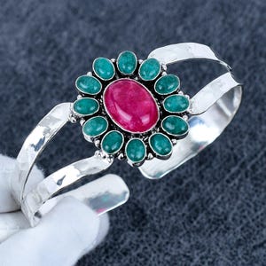 Op de afbeelding: Een zilveren manchetarmband met een centrale ovale, dieproze edelsteen, omringd door kleinere, ovale, teal-kleurige stenen. De armband heeft een gehamerde textuur en een gespleten ontwerp.