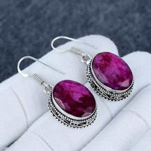 Peut inclure: Une paire de boucles d'oreilles pendantes argentées. Chaque boucle d'oreille présente une grosse pierre précieuse rose foncé de forme ovale, sertie dans une lunette décorative. Les boucles d'oreilles ont une fermeture à crochet et un élément de conception en spirale.
