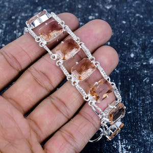 Op de afbeelding: Een zilveren armband met rechthoekige, perzik-kleurige edelstenen. De armband bestaat uit onderling verbonden schakels met kleine, heldere accentstenen. De edelstenen zijn gezet in een zilveren metalen frame.