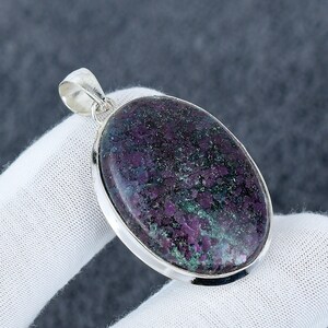 Puede incluir: Colgante ovalado con una piedra pulida de color púrpura oscuro y verde engastada en un marco de metal plateado. La piedra tiene un aspecto moteado. El colgante tiene un lazo en la parte superior para una cadena.