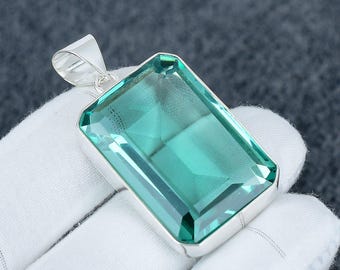 May include: A rectangular, teal-colored gemstone pendant with a silver bail. The gemstone is set in a silver bezel and has a faceted surface. The pendant is displayed on a white surface.