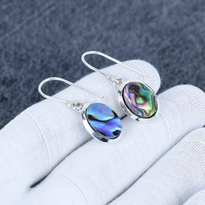Può includere: Orecchini in argento con conchiglie di abalone ovali. I gusci iridescenti mostrano un mix di tonalità blu, verdi e viola. Gli orecchini sono incastonati in una lunetta d'argento e hanno un semplice design a gancio.
