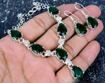 May include: A silver necklace and matching earrings set featuring pear-shaped, dark green gemstones. The necklace has a delicate chain and floral accents. The earrings have a simple hook design. The jewelry is displayed on a hand.