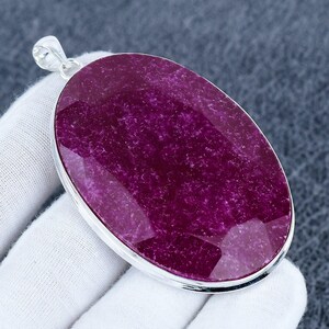May include: An oval pendant featuring a deep magenta gemstone set in a silver frame. The gemstone has a faceted surface, reflecting light. The pendant is suspended from a silver bail. The pendant is likely made of silver.